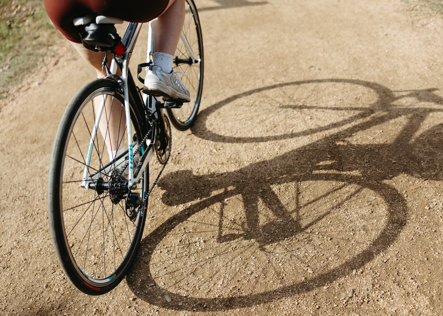 Close-up of a person riding a bicycle on a dirt path near UMich Off-Campus Housing, showing the back wheel, part of their legs, and the bike's shadow cast on the ground.