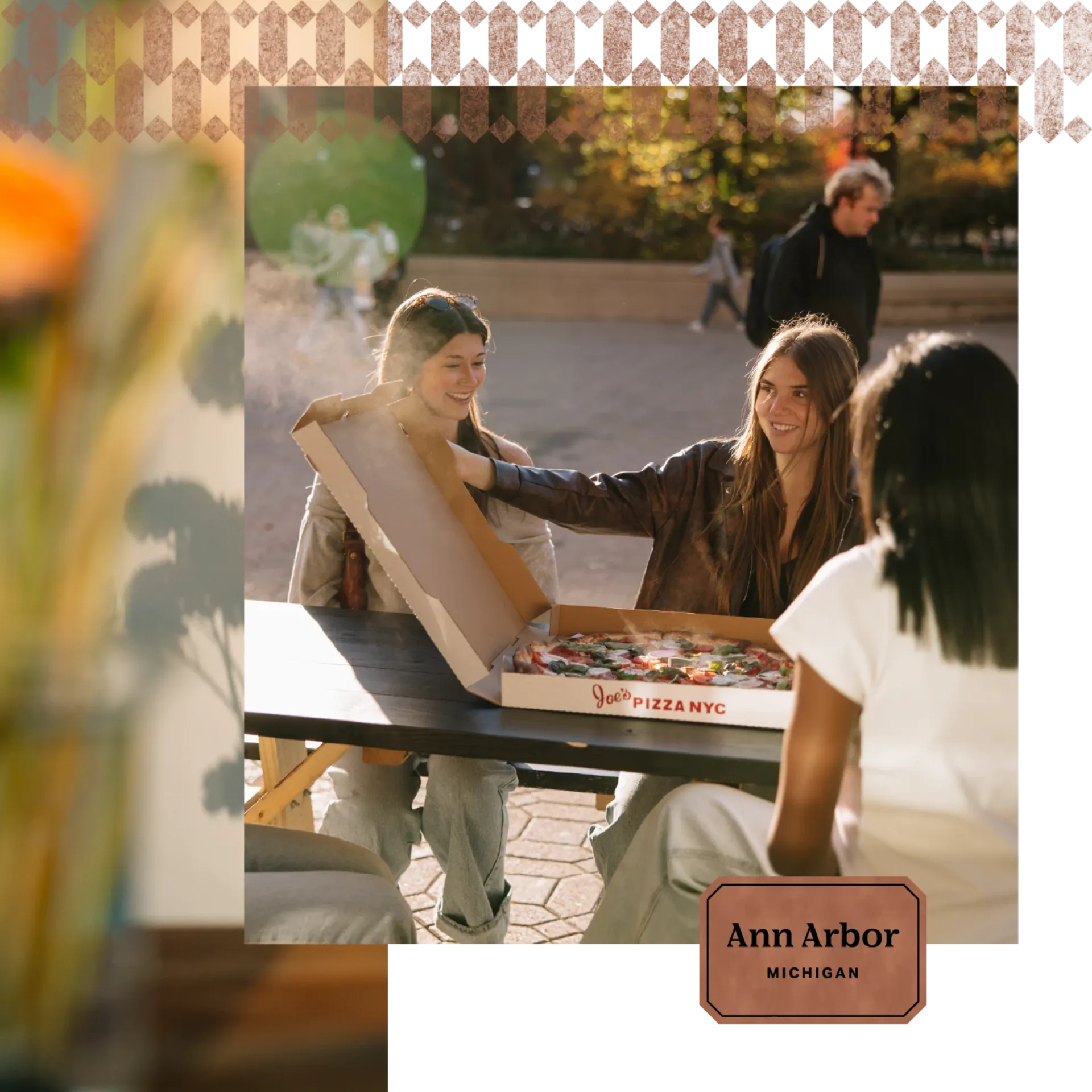 Three young women sit at an outdoor table, smiling and opening a pizza box together in Ann Arbor. Sunlight and steam rise from the pizza, capturing the vibrant atmosphere near Ann Arbor Student Apartments.