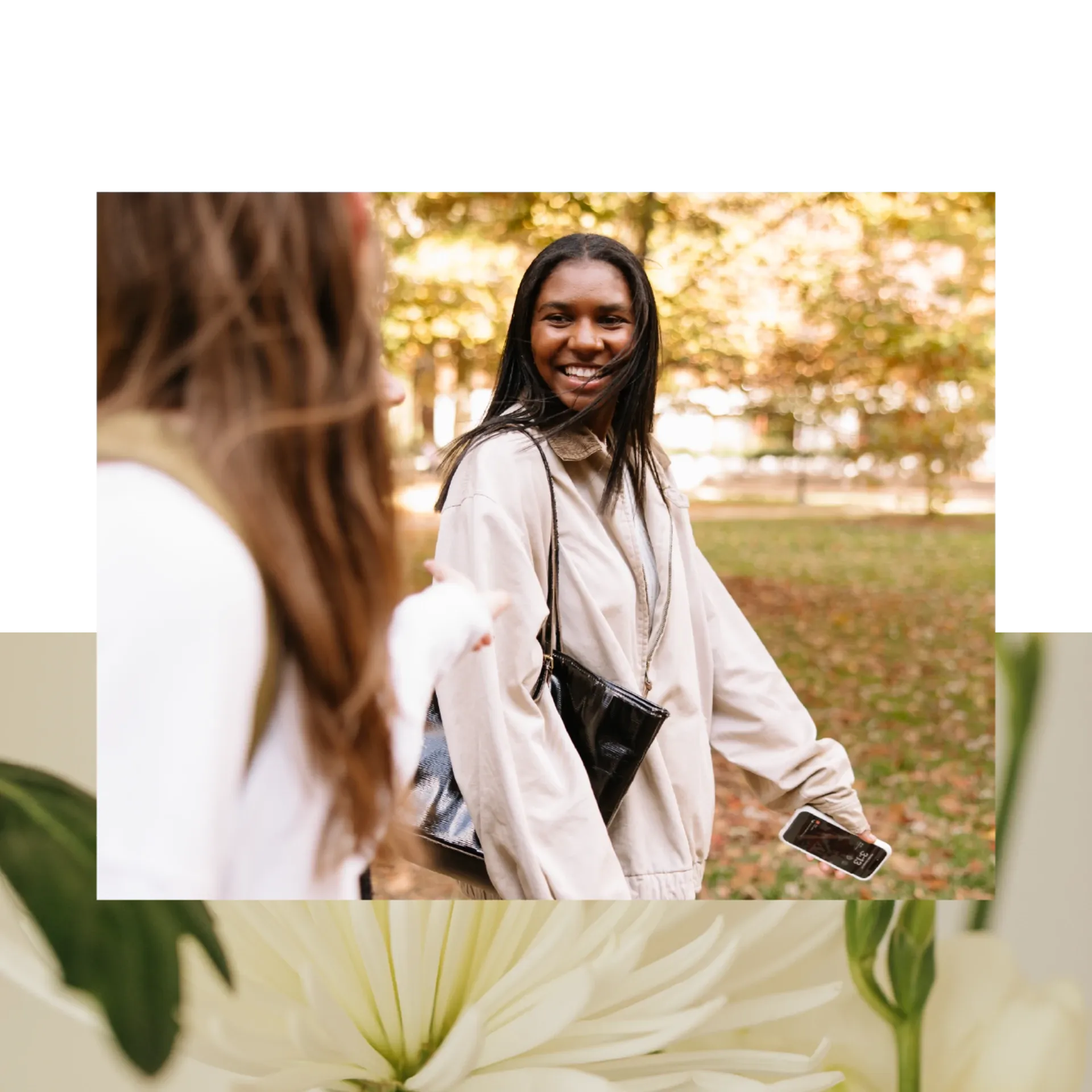 Two women walk outdoors in a park with autumn leaves; one, smiling, looks back over her shoulder while holding a phone and a black purse—perhaps exploring the neighborhood near Ann Arbor Student Apartments. Blurred flowers appear at the bottom.