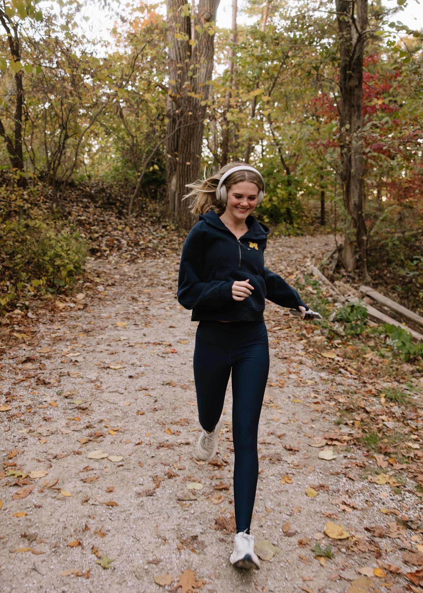 A woman wearing headphones, a dark hoodie, and leggings jogs on a leaf-strewn forest trail in autumn near Ann Arbor Student Apartments, smiling as she runs.