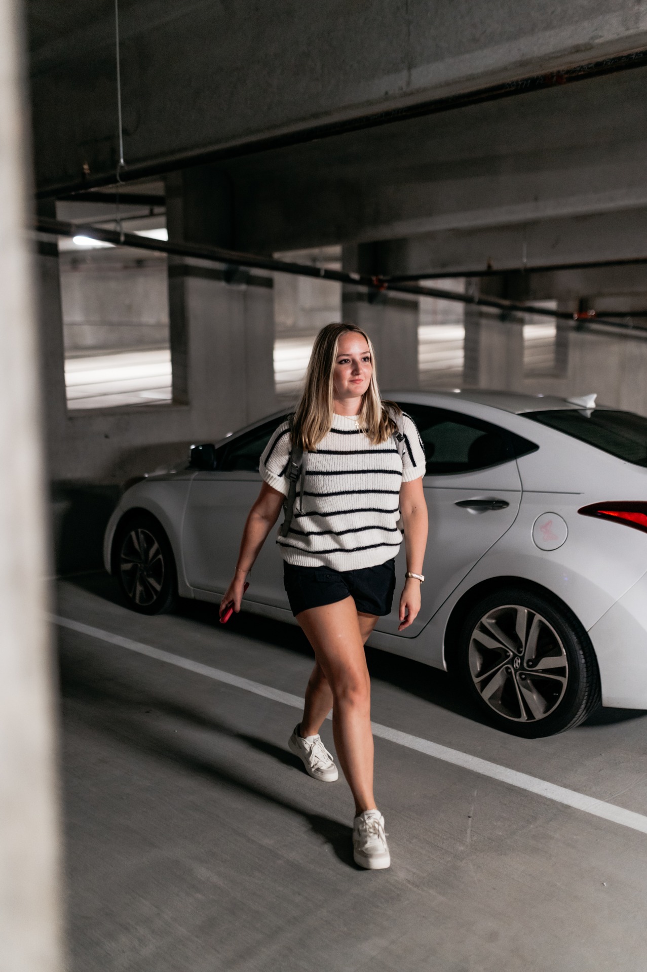 A woman wearing a striped shirt, black shorts, and white sneakers walks confidently in a UMich Off-Campus Housing parking garage beside a white car, holding a phone in her right hand.