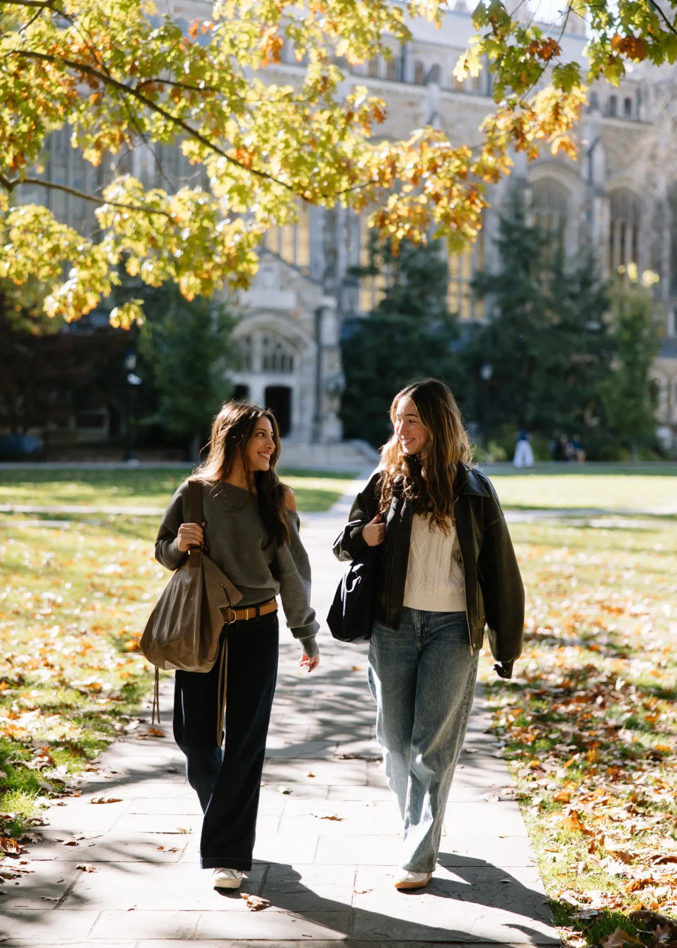 Two young women walk together on a tree-lined campus path near UMich Off-Campus Housing, smiling and talking. They carry bags and wear casual clothes. Autumn leaves are scattered on the ground, with a large building in the background.
