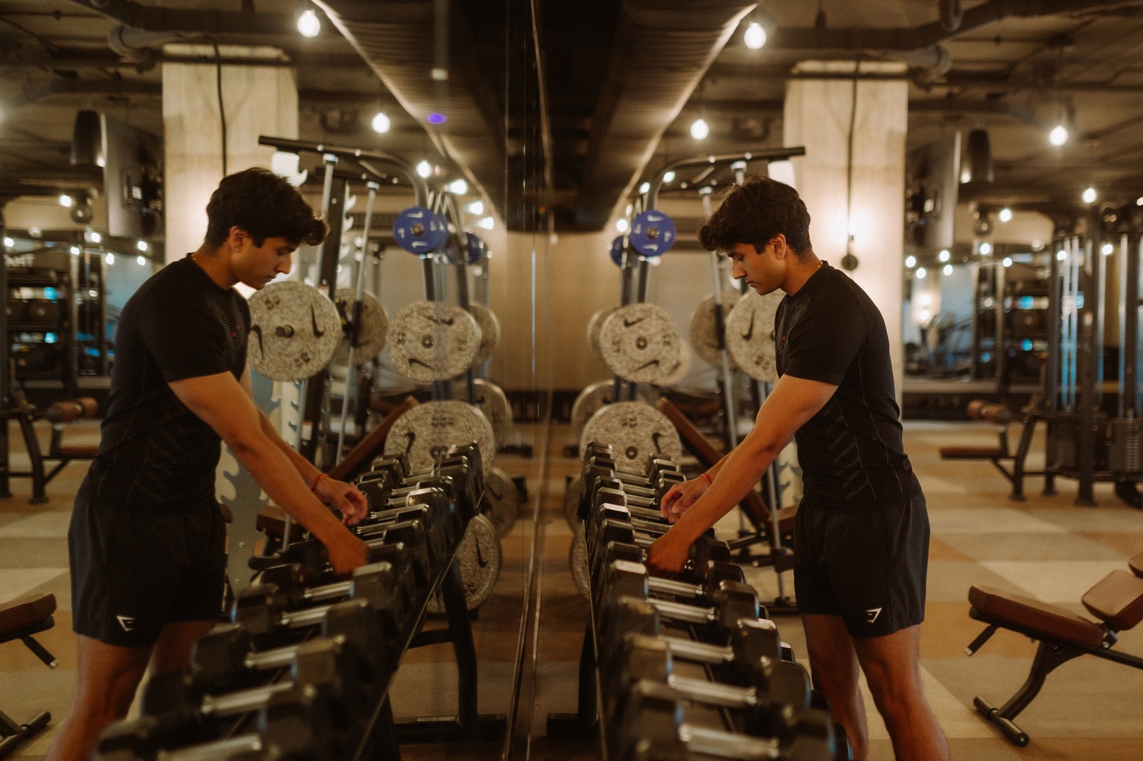 A man in athletic wear stands beside a rack of dumbbells in a gym, located within UMich Off-Campus Housing, looking down as he selects a weight. His reflection is visible in the mirror amidst well-lit, modern exercise equipment.