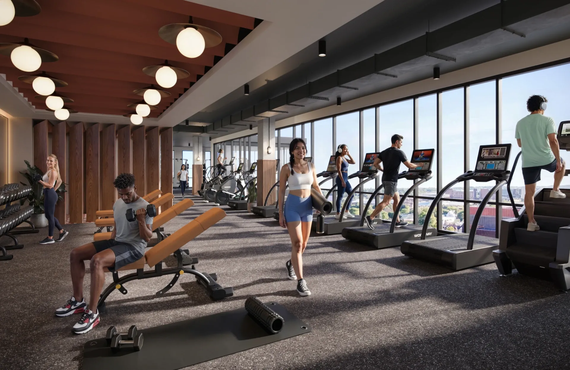 A modern gym in UMich Off-Campus Housing features people using treadmills, lifting weights, stretching, and exercising. Natural light fills the room, energizing the bright space with city views just outside.