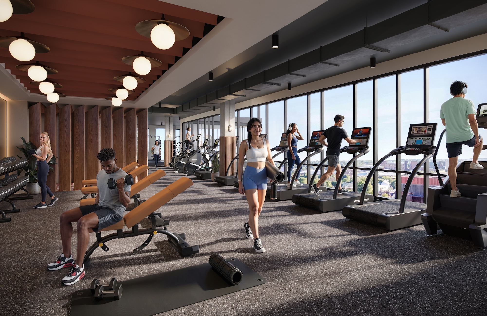 People work out in a modern gym with large windows, surrounded by UMich Off-Campus Housing, using treadmills and dumbbells. Natural light fills the space, and the city skyline is visible outside.