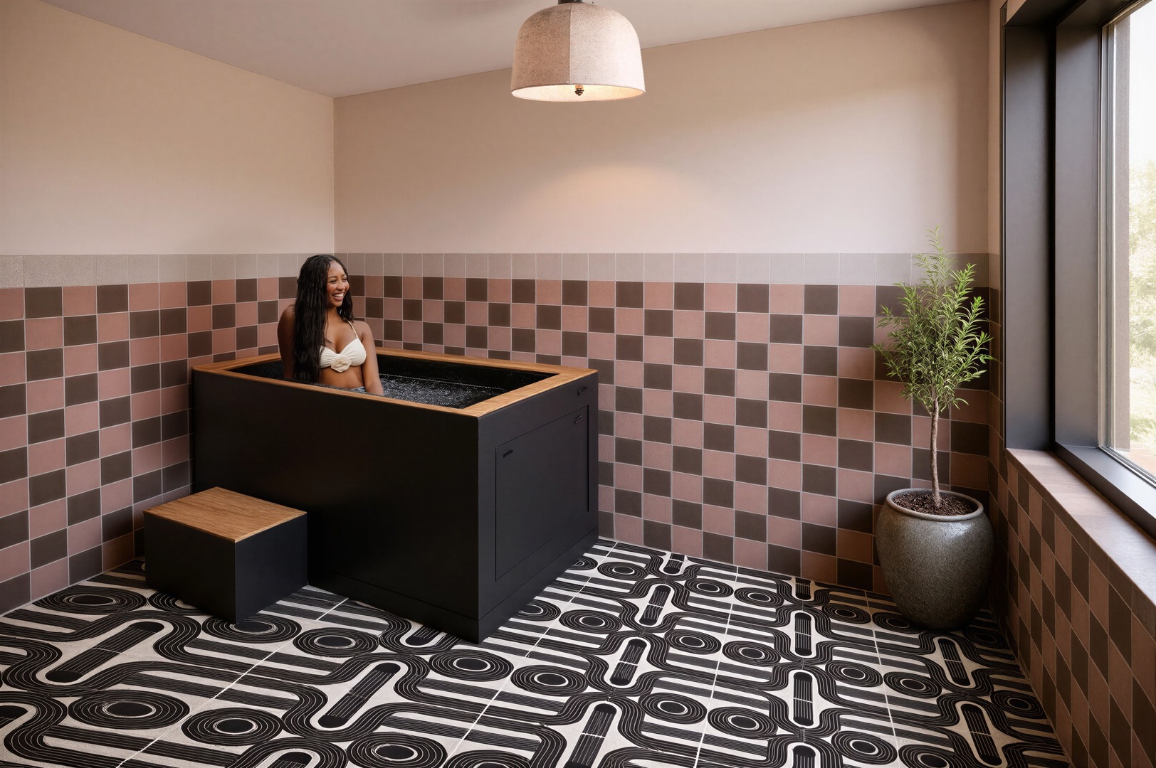 A woman in a white bikini top sits in a modern black soaking tub in a stylish UMich Off-Campus Housing bathroom with patterned floors, brown checkered walls, a potted plant, and a large window letting in natural light.