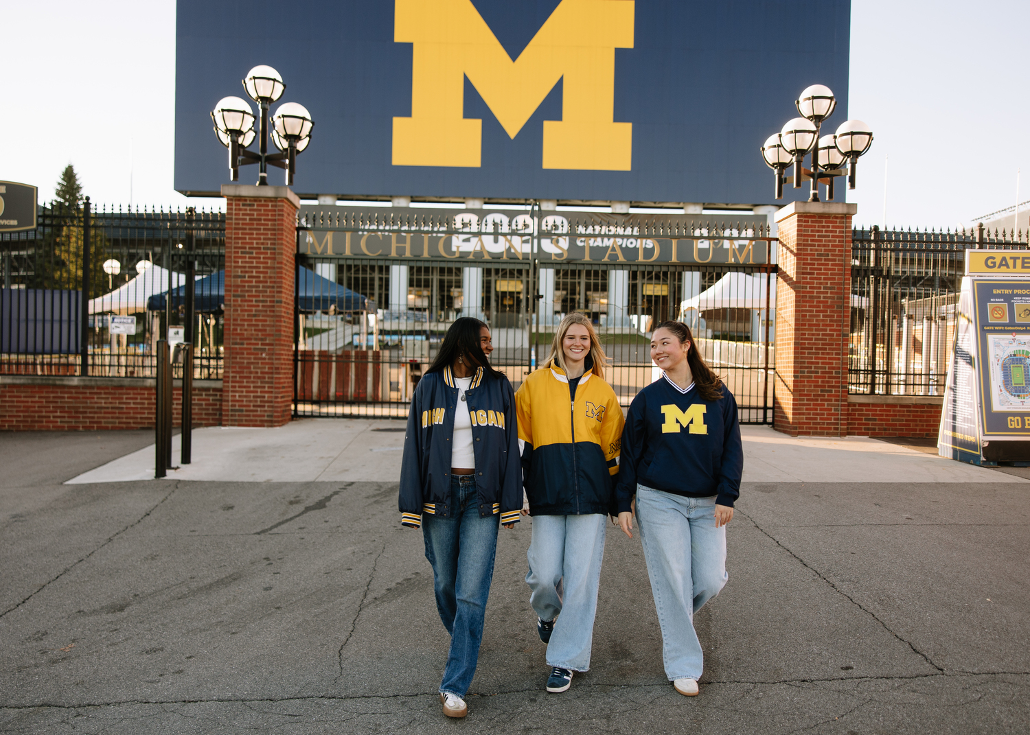 Three women wearing University of Michigan apparel walk together and smile in front of Michigan Stadium, with a large maize and blue M sign visible behind them, perhaps chatting about the cost to live in an Ann Arbor apartment.