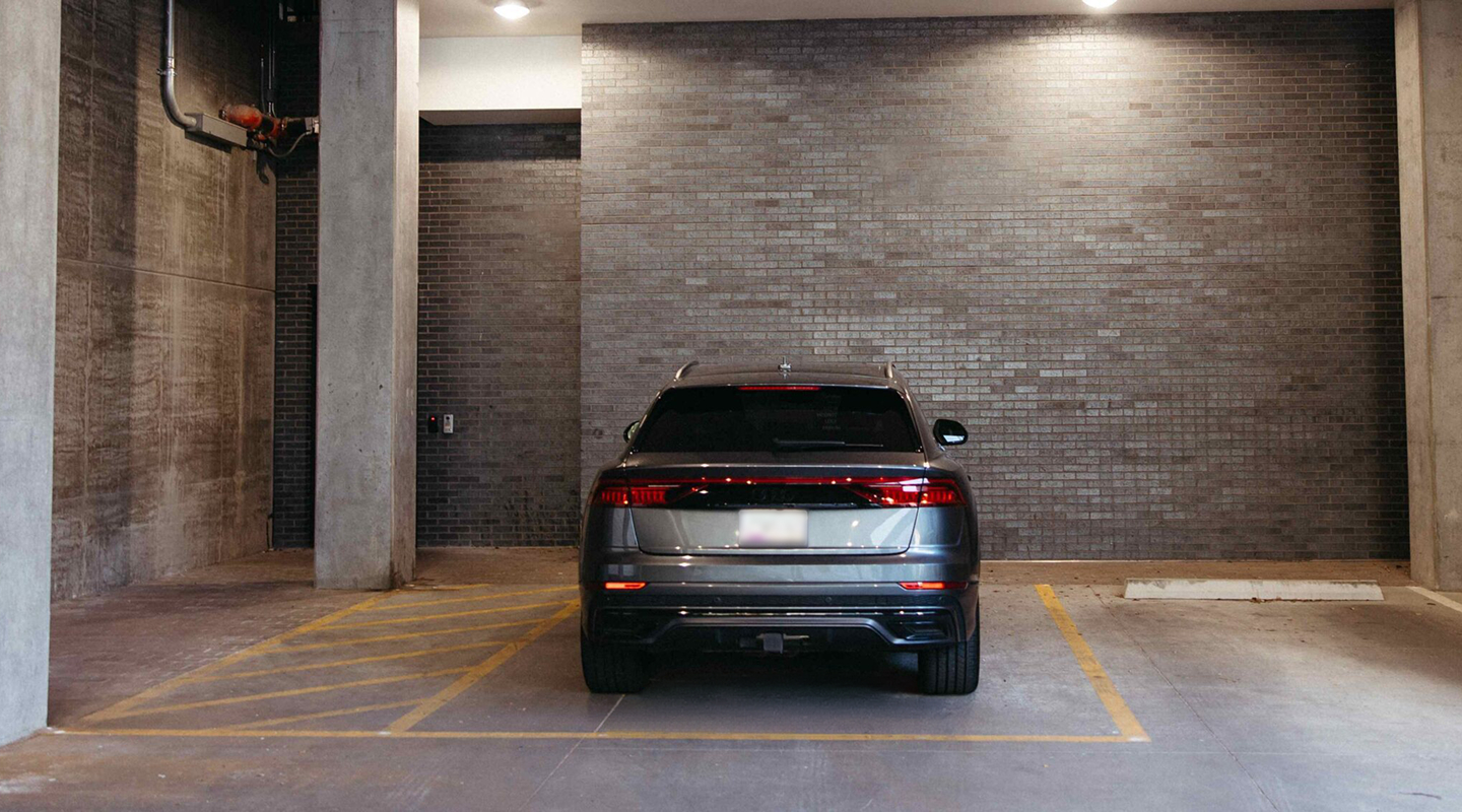 A gray SUV is parked in front of a brick wall inside a parking garage, occupying a space next to an empty, yellow-striped accessible zone near UMich Off-Campus Housing.
