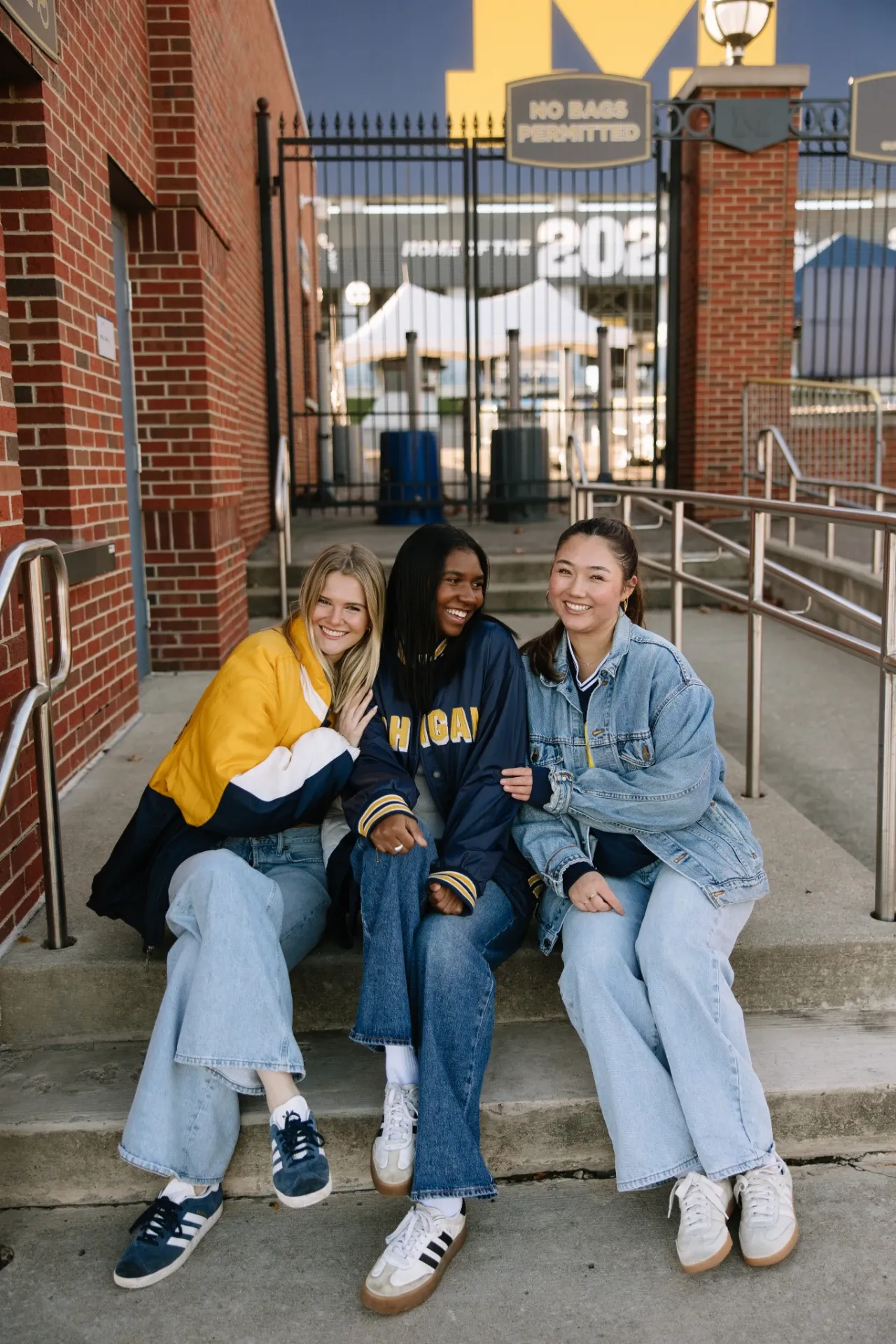 Three young women sit together on steps outside a stadium gate, smiling. They wear casual outfits including jeans, sneakers, and jackets, with University of Michigan apparel visible.