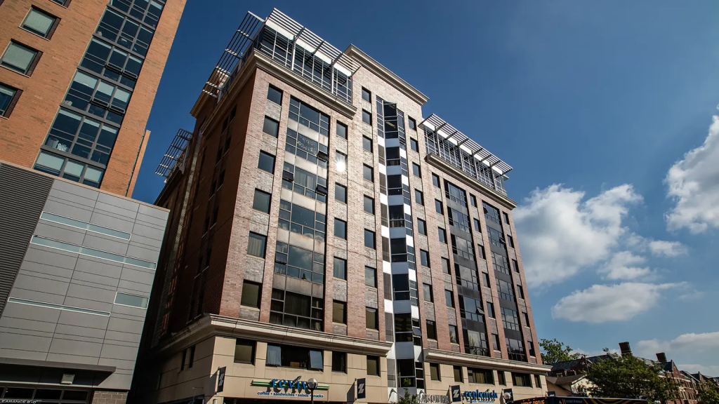 A modern mid-rise building offering new student apartments, featuring large windows, a brick and beige facade, and retail stores on the ground floor, set against a blue sky with scattered clouds.