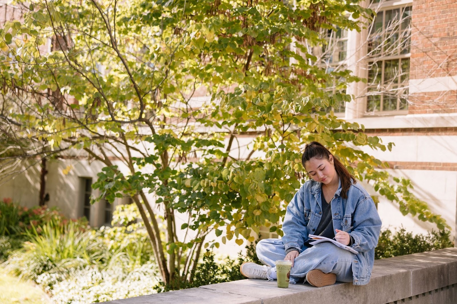 A young woman in a denim jacket and jeans sits cross-legged on a concrete ledge outdoors, signing a lease in her notebook with a drink beside her. Green trees and a brick building are in the background.