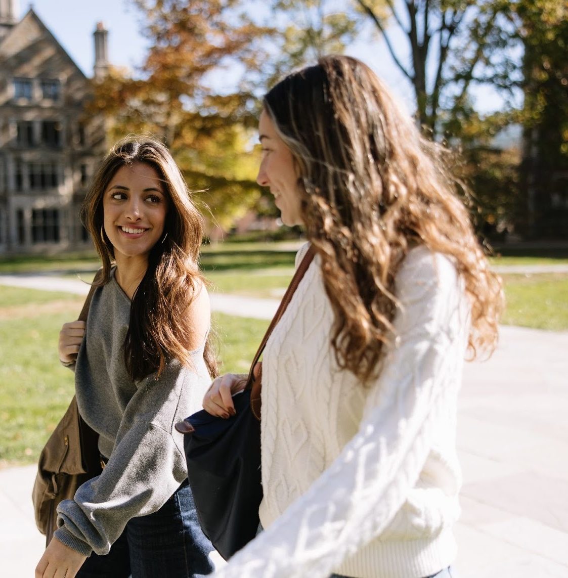 Two young women, college roommates, carrying bags, walk and smile at each other on a university campus with historic stone buildings and autumn trees in the background.