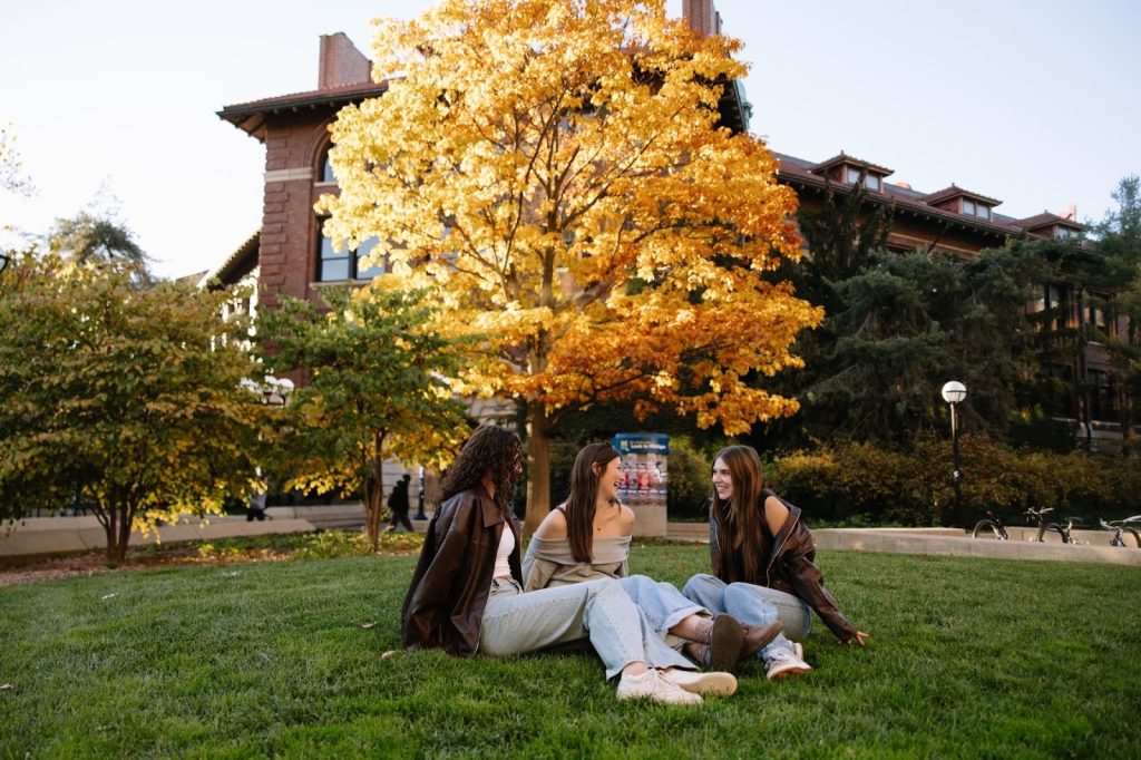 Three UMich students sitting on the lawn under colorful fall trees near campus in Ann Arbor.