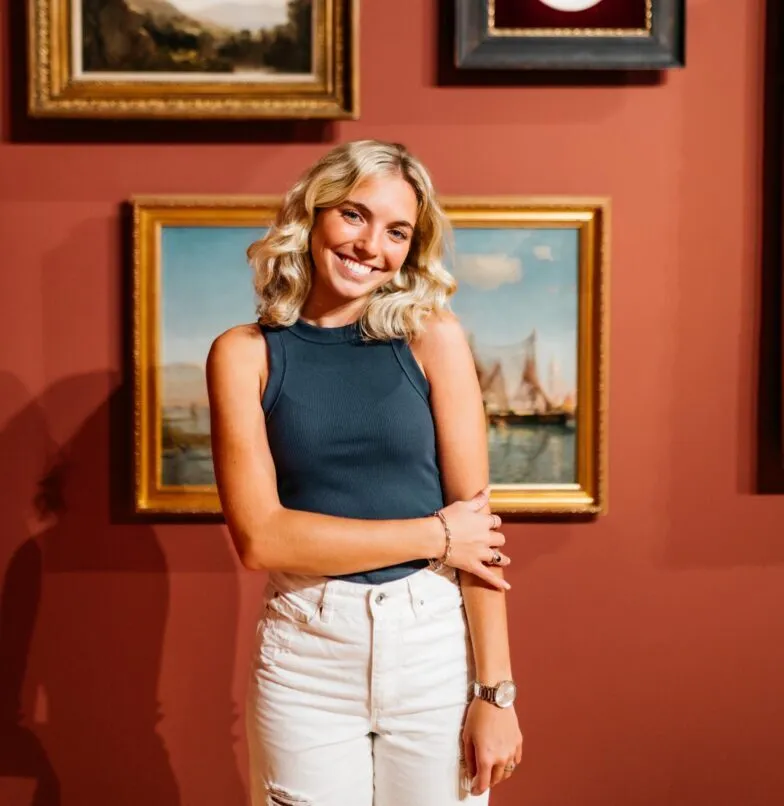 A smiling woman with blonde hair, wearing a sleeveless dark top and white pants, stands in front of framed paintings on a reddish wall in a gallery.