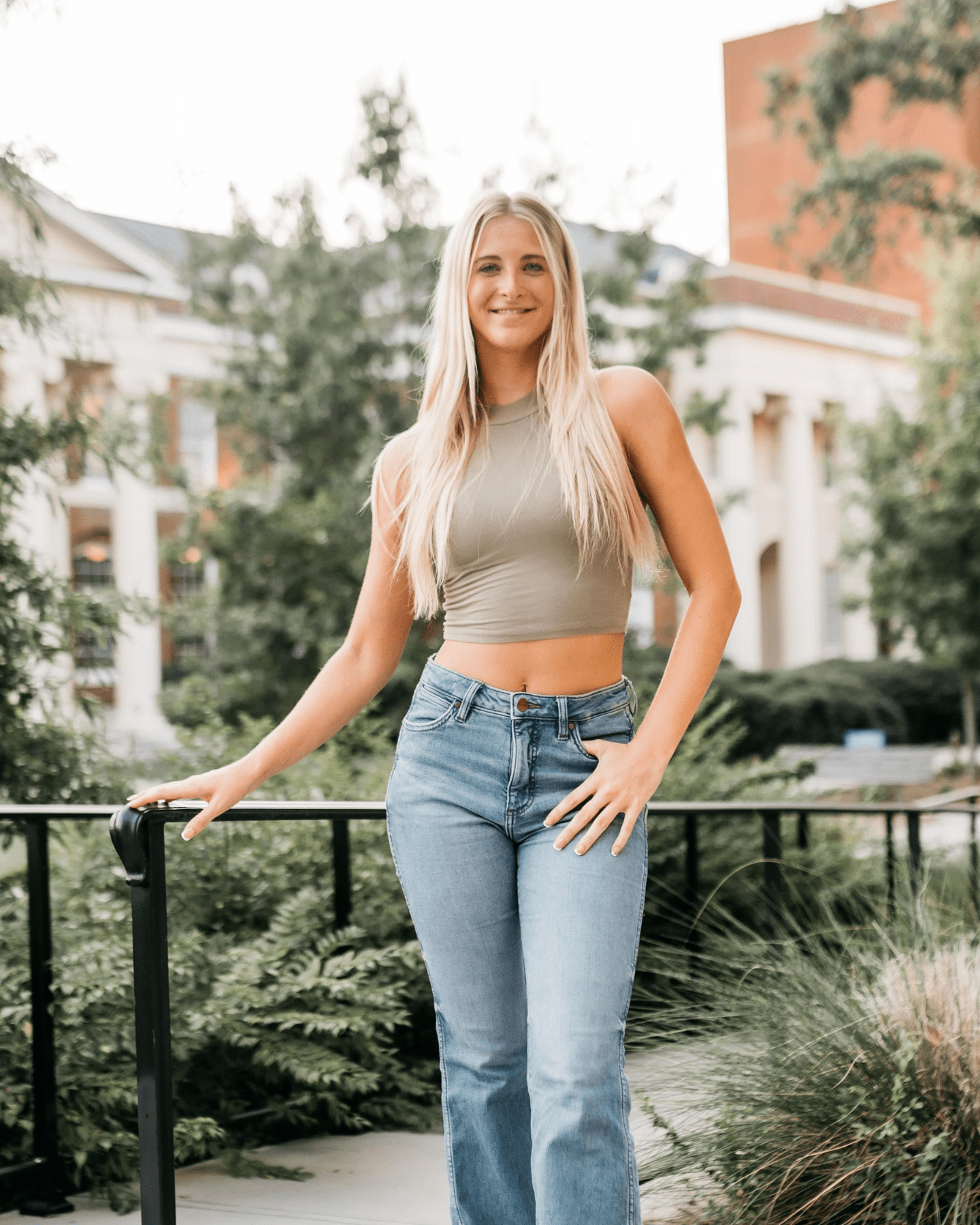 A young woman with long blonde hair, wearing a sleeveless olive crop top and blue jeans, stands outdoors by a black railing, with greenery and a building in the background.