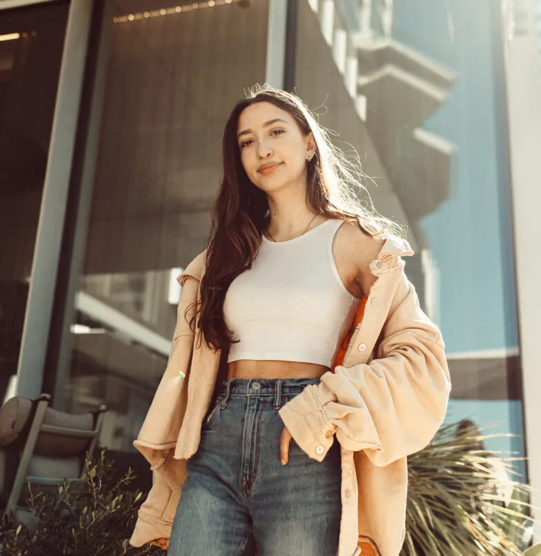 A young woman with long brown hair stands outside a modern glass building, wearing a white crop top, high-waisted jeans, and an open tan jacket. She looks confidently at the camera with a slight smile.