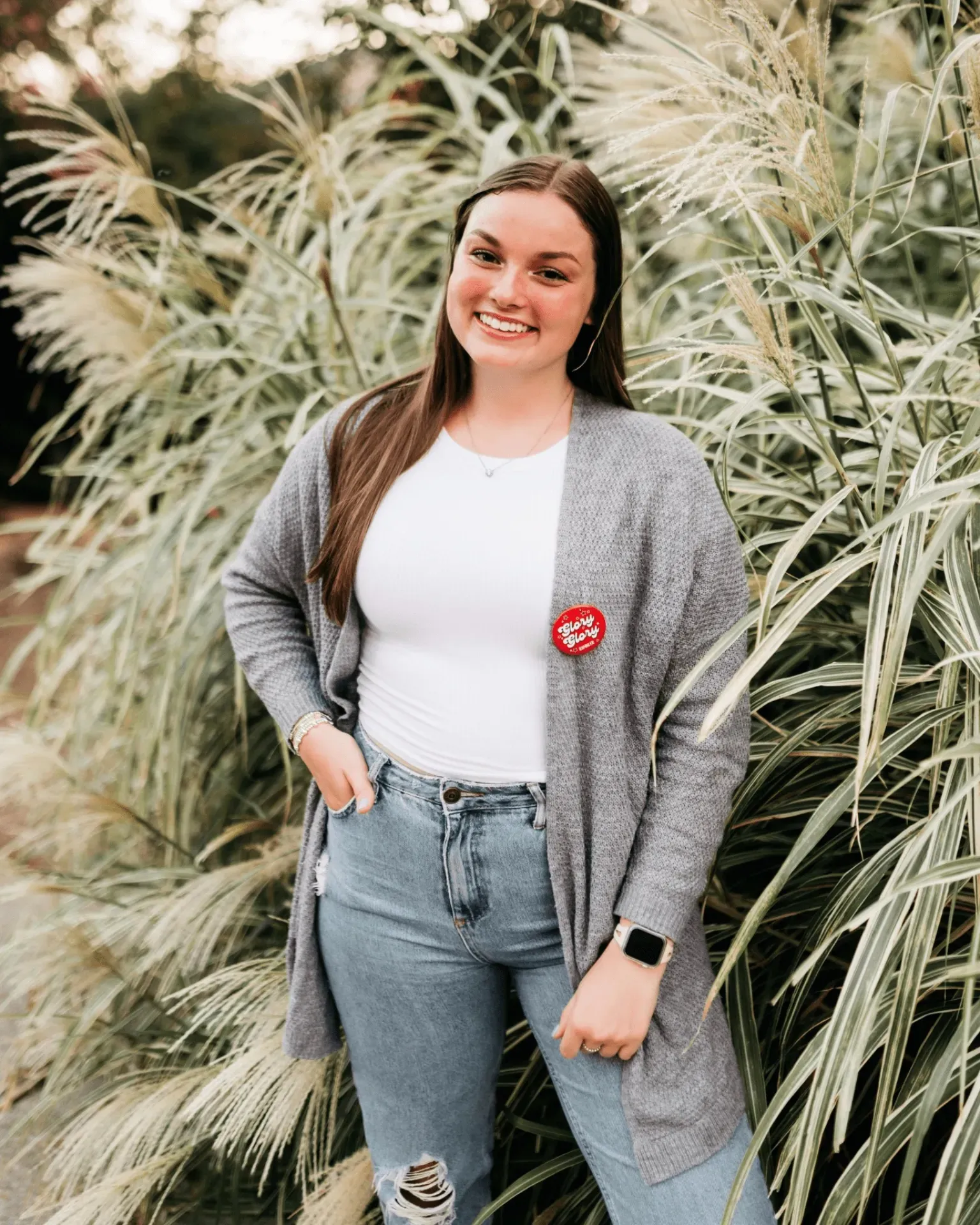 A smiling young woman stands outdoors in front of tall, green grasses. She wears a gray cardigan, white shirt, ripped blue jeans, and a red “I Voted” sticker on her shirt. Her hands rest on her hips.