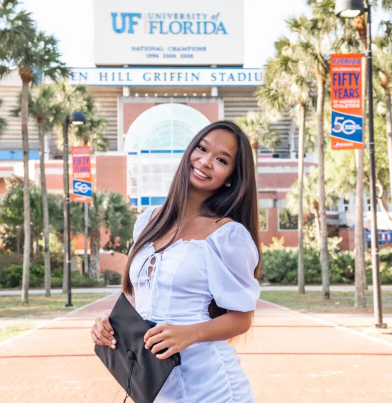 A young woman in a white dress holds a graduation cap and smiles in front of the University of Florida’s Ben Hill Griffin Stadium, with palm trees and anniversary banners in the background.