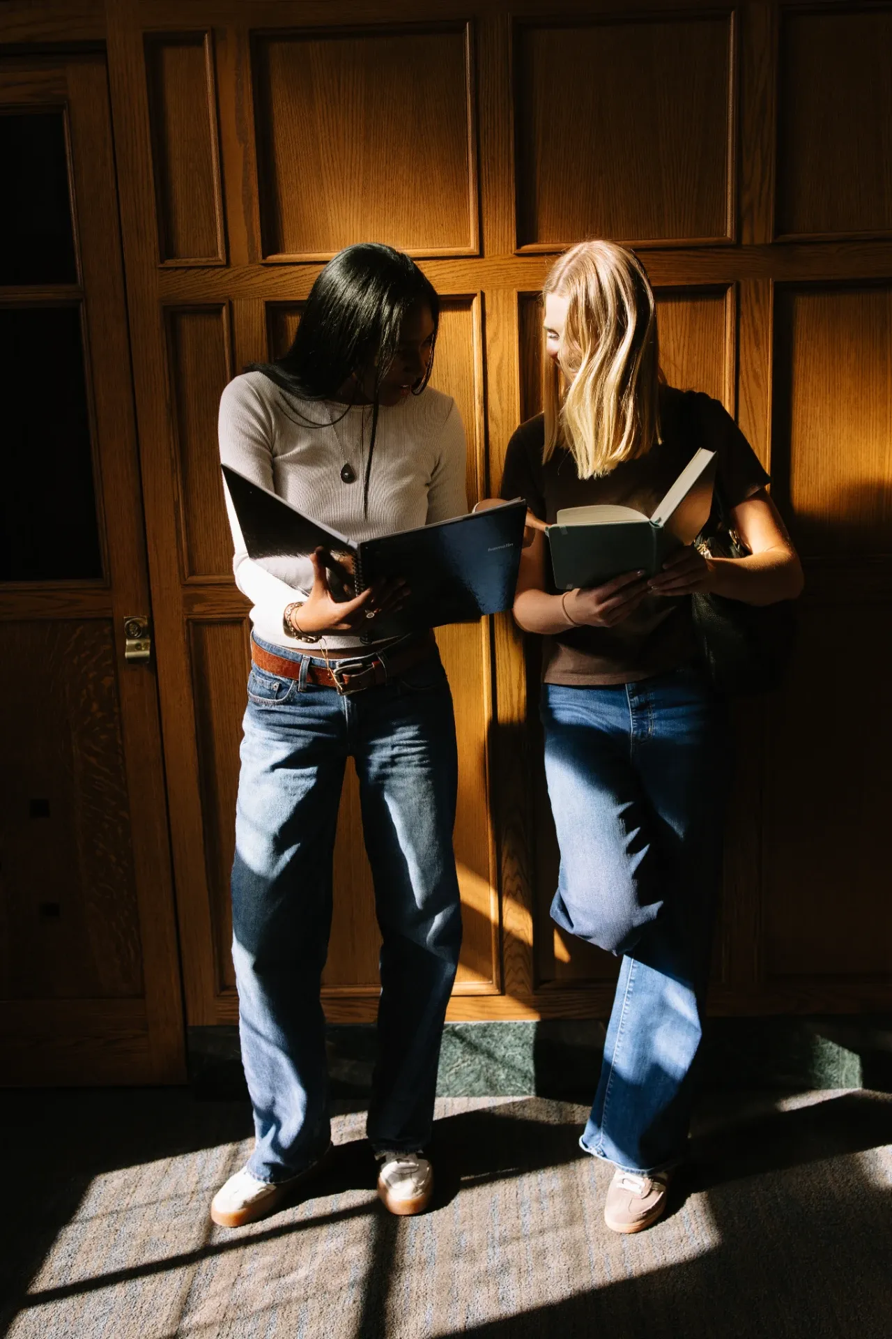 Two young women stand side by side against a wooden wall, reading books. They are dressed casually in jeans and long-sleeve shirts, with sunlight streaming in and casting shadows around them.