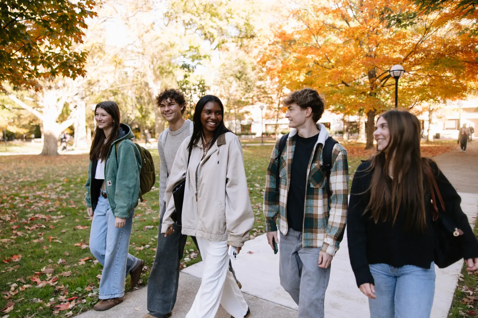 Five young adults walk together outdoors on a college campus in autumn, smiling and talking. Colorful fall leaves are visible on the trees, and they wear casual clothes and backpacks.