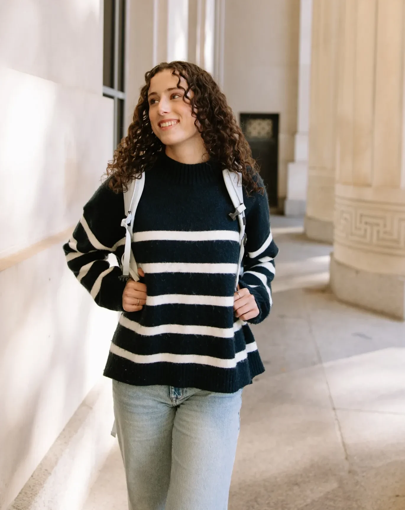 A person with curly hair, wearing a black and white striped sweater, light jeans, and a backpack, walks outside near large columns, smiling and looking to the side.