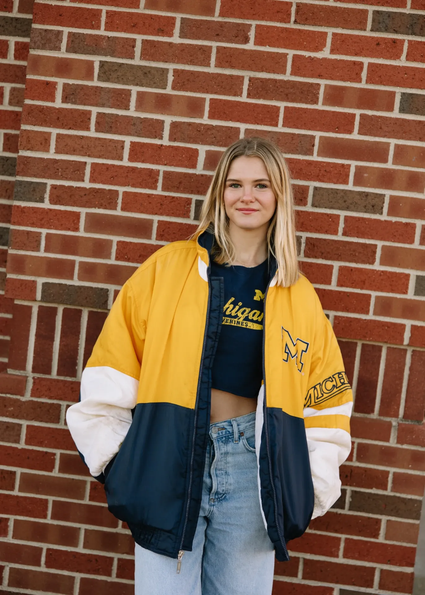 A young woman stands in front of a brick wall, wearing a yellow and blue Michigan jacket over a matching T-shirt and blue jeans, with her hands in her pockets and a slight smile on her face.