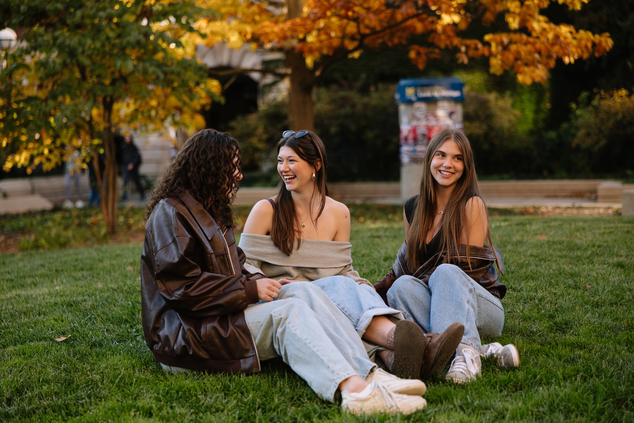 Three young women sit on grass near Michigan off campus housing, laughing and talking together. They wear casual clothes, with autumn trees of orange leaves behind them. The mood is relaxed and cheerful.