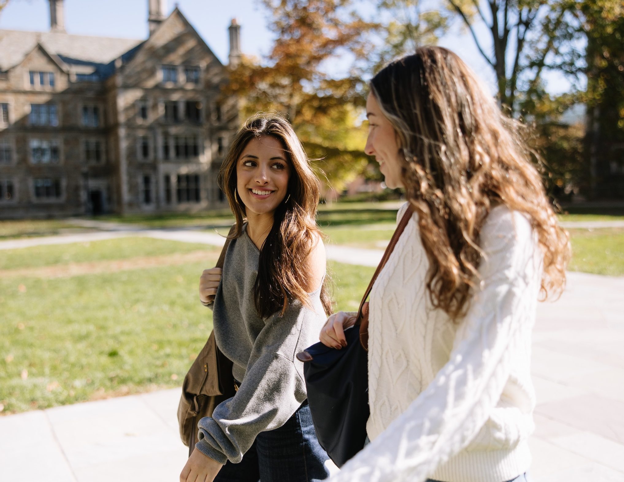 Two young women walk outside on a sunny day, smiling and talking near a large stone building and green lawn. Both are carrying bags and dressed casually—typical of friends heading to or from their nearby UMich student apartments.