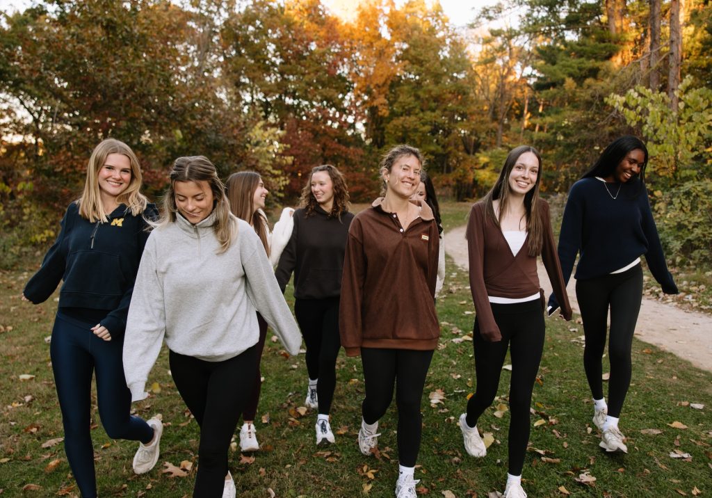 Seven young women walk together on a path in a park surrounded by autumn trees, smiling and enjoying each other's company. Dressed casually, they chat about life in their umich student apartments and savor the crisp fall air.
