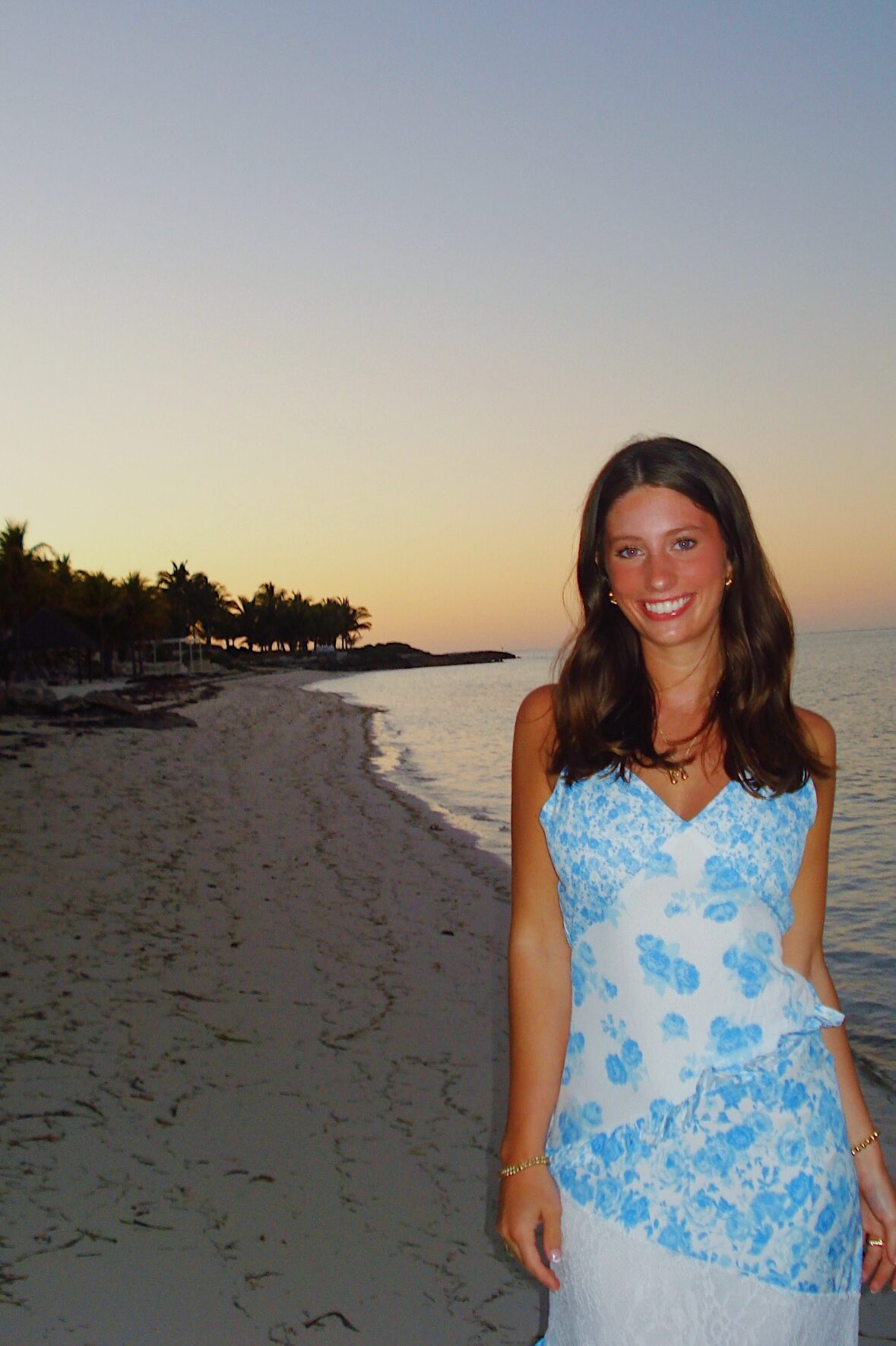 A woman in a blue and white floral dress stands on a sandy beach at sunset, smiling at the camera—her carefree vibe echoes the relaxed lifestyle found in umich student apartments, with calm water and palm trees in the background.
