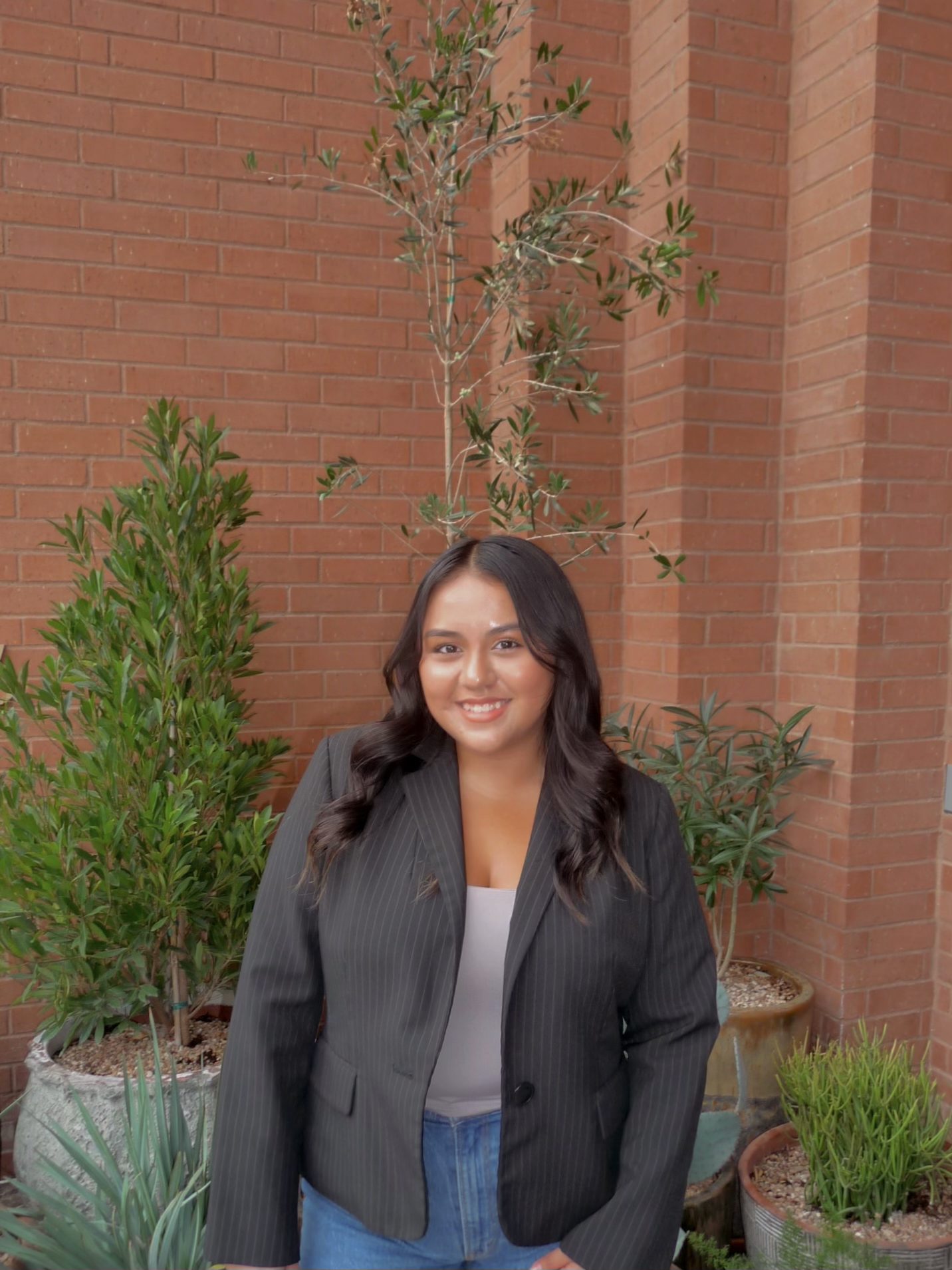 A woman with long dark hair, wearing a black pinstripe blazer and blue jeans, stands smiling in front of a brick wall with potted green plants around her—capturing the welcoming vibe of umich student apartments.