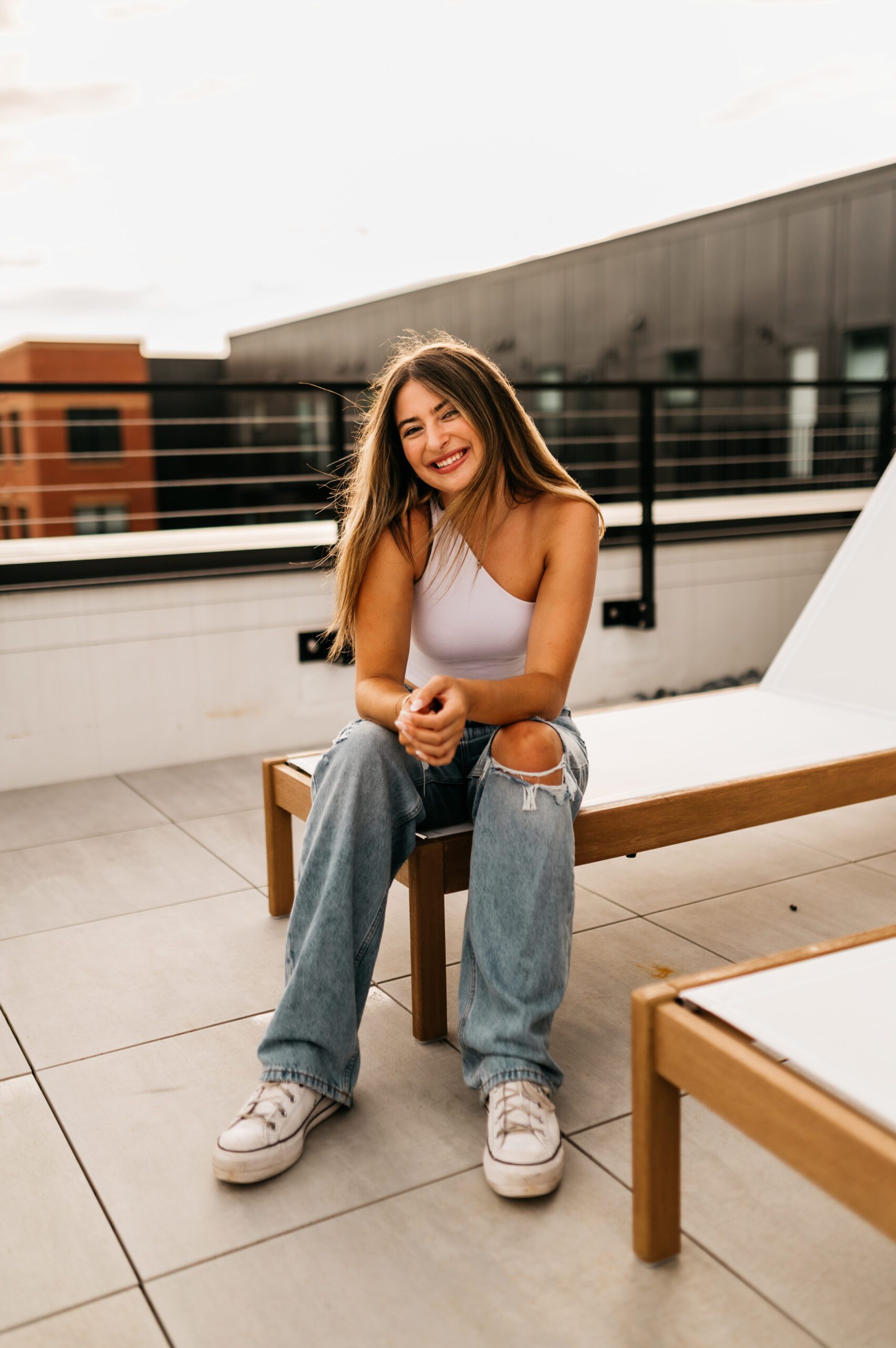 A young woman with long hair, wearing a white one-shoulder top, ripped jeans, and white sneakers, sits smiling on a wooden lounge chair atop the rooftop patio of stylish UMich student apartments, with modern buildings in the background.