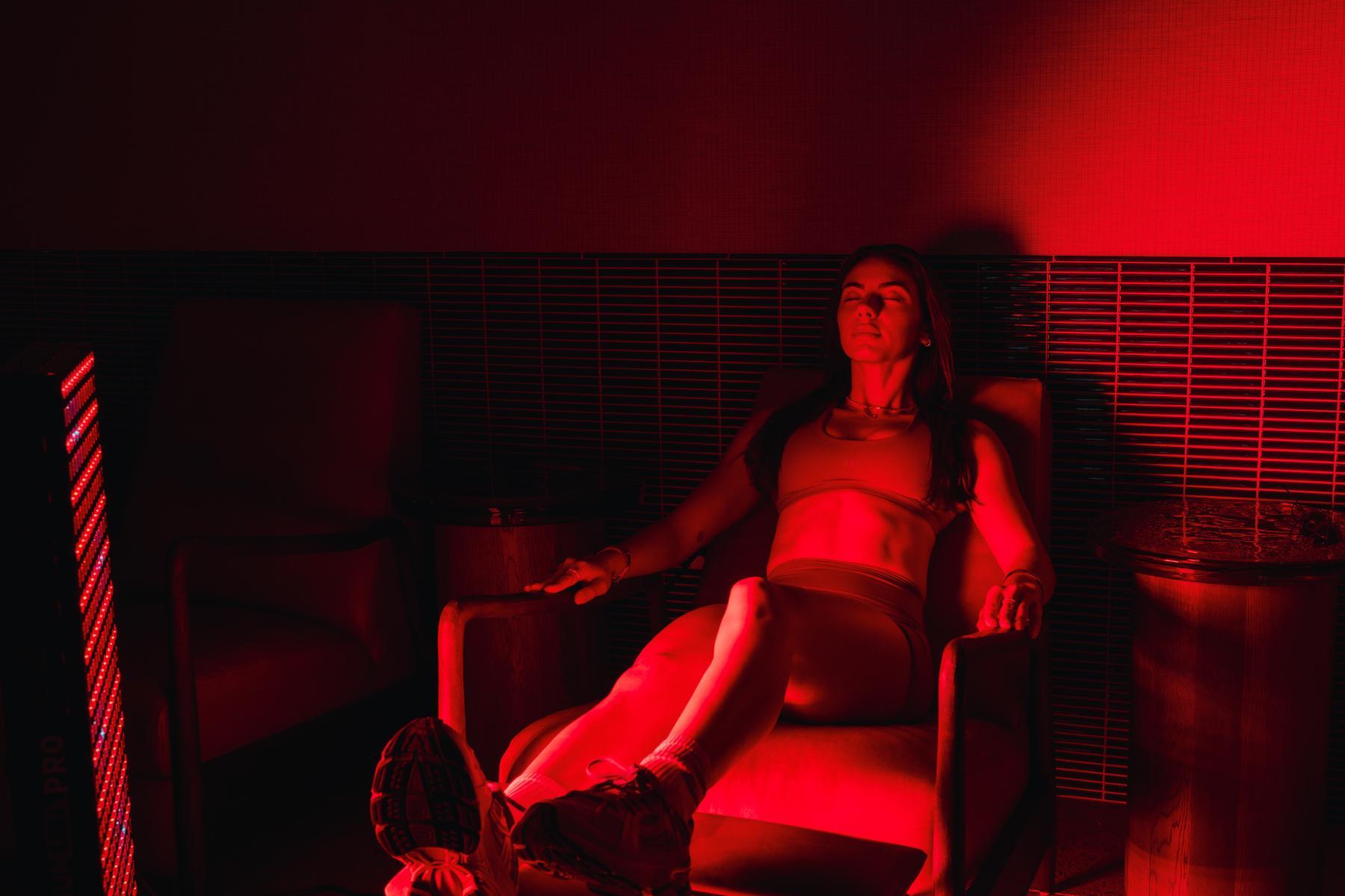 A woman in athletic wear sits relaxed in a chair, bathed in intense red light, with her eyes closed and arms resting on the armrests—a calm moment after a long day exploring UMich Off-Campus Housing options.