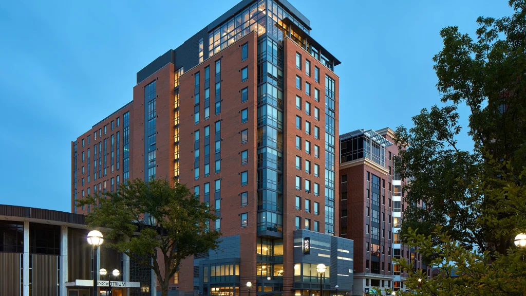 Six11 student apartment building in Ann Arbor, a modern red-brick high-rise with large windows and a glass corner façade, located near the University of Michigan campus at dusk.