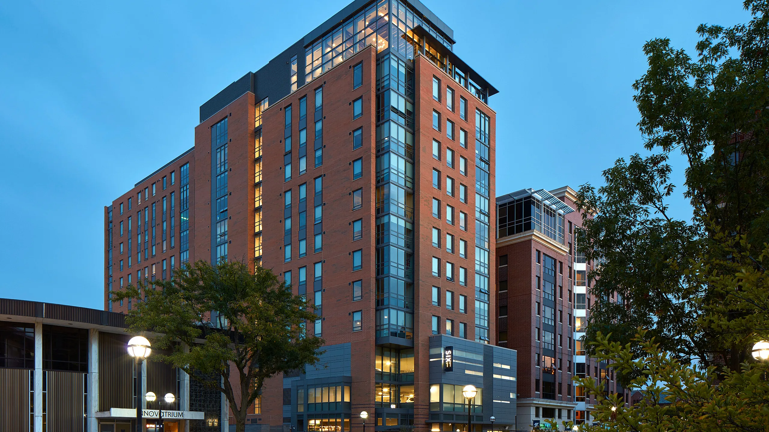 Six11 student apartment building in Ann Arbor, a modern red-brick high-rise with large windows and a glass corner façade, located near the University of Michigan campus at dusk.