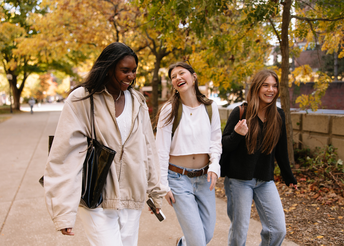 Three young women walk outside on a tree-lined sidewalk in autumn, smiling and laughing together as they head to their UMich Off-Campus Housing, enjoying a pleasant day in casual clothes and carrying bags.