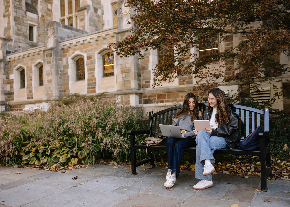 Two young women sit on a bench outside a stone building in verve ann arbor, smiling as they work on a laptop and tablet. Autumn leaves and greenery surround them, creating a relaxed academic atmosphere.