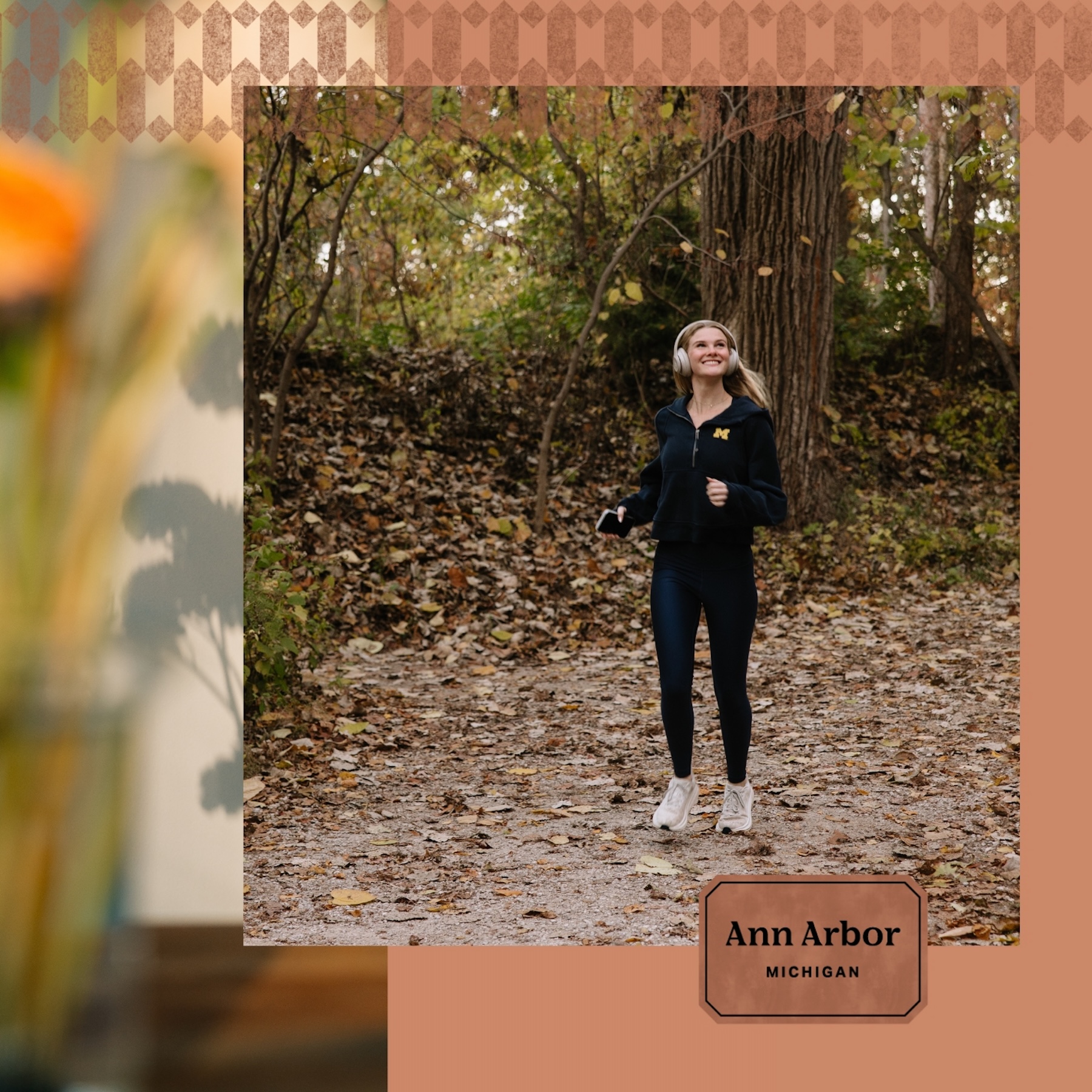 A woman in athletic wear jogs on a leaf-covered trail in a wooded area during autumn, near UMich student apartments with wellness amenities. A text box in the corner reads Ann Arbor, Michigan.