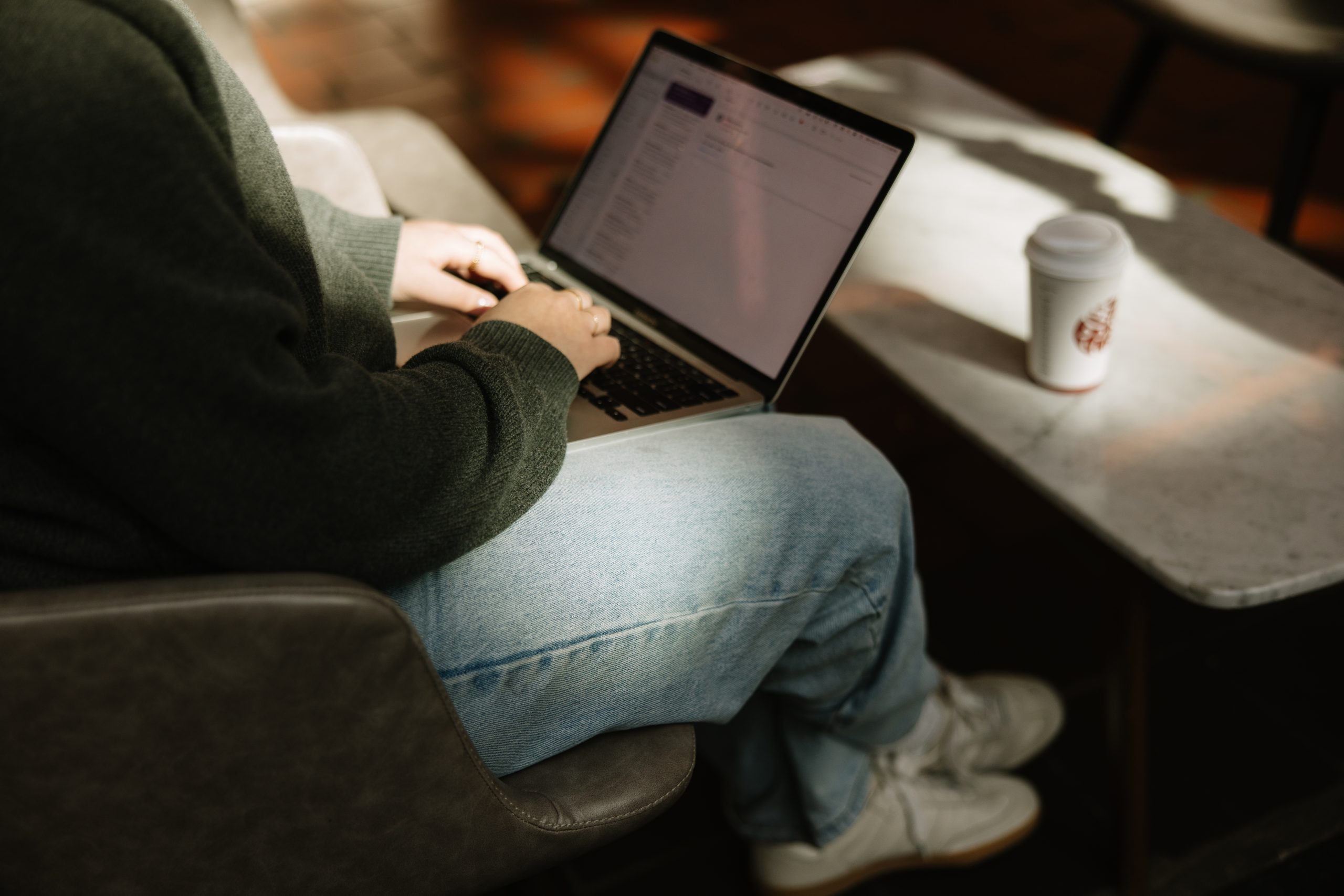 A person sitting in an armchair working on a laptop with a coffee cup on the table beside them.